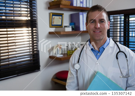 Male doctor standing in clinic holding blue folder wearing lab coat with stethoscope, copy space 135307142