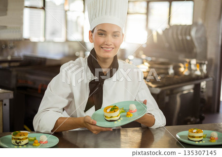 Female chef in uniform plating layered desserts with fruit garnish on counter in kitchen with ovens Female chef in uniform plating layered desserts with fruit garnish on counter in kitchen with ovens 135307145