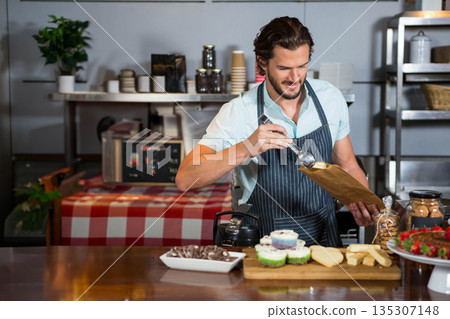 Man scooping cupcakes with metal scoop into paper bag on checkered cloth at bakery counter Man scooping cupcakes with metal scoop into paper bag on checkered cloth at bakery counter 135307148
