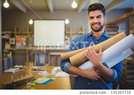 Man holding rolled plans while smiling and standing in studio with worktable and models, copy space 135307150