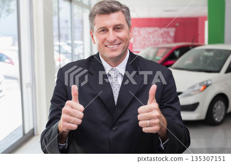 Man wearing business suit standing in car showroom holding thumbs up near display cars and windows 135307151