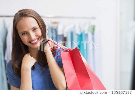 Woman in her twenties smiling while holding red shopping bags by store clothing rack, copy space 135307152