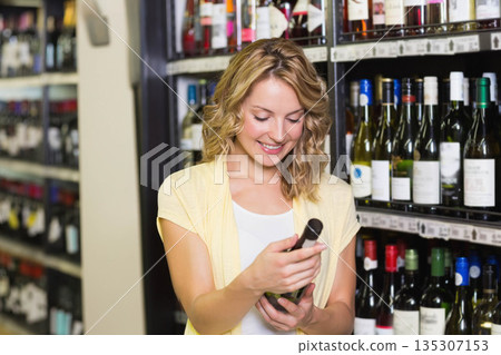 Female shopper holding wine bottle and reading label in retail wine aisle with metal shelves Female shopper holding wine bottle and reading label in retail wine aisle with metal shelves 135307153