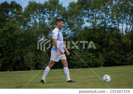 Male soccer player walking across grass field dribbling soccer ball wearing blue jersey and cleats 135307159