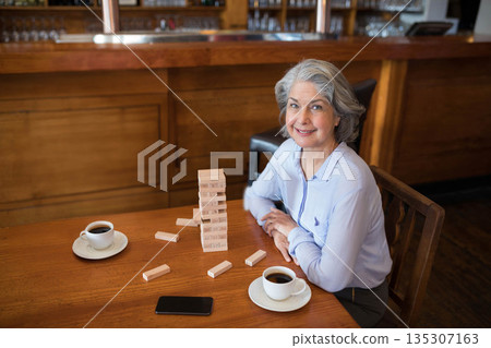 Senior woman sitting in cafe building block tower with coffee cups and smartphone smiling at camera Senior woman sitting in cafe building block tower with coffee cups and smartphone smiling at camera 135307163