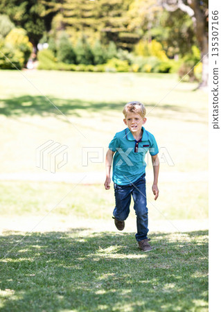 Boy running across sunlit grassy park field wearing turquoise polo shirt and dark jeans 135307166