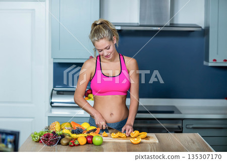 Woman wearing sportswear slicing oranges with chef's knife on wooden cutting board in home kitchen 135307170