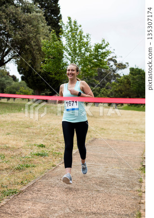 Woman wearing smartwatch and bib number 1671, breaking red finish line tape on paved park path 135307174