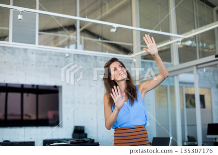 Woman gesturing toward virtual interface on glass partition in open-plan office with wall monitor Woman gesturing toward virtual interface on glass partition in open-plan office with wall monitor 135307176