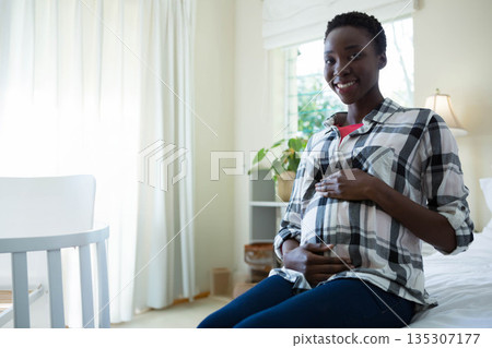 African American woman holding pregnant belly sitting on bed in nursery with white crib, copy space African American woman holding pregnant belly sitting on bed in nursery with white crib, copy space 135307177