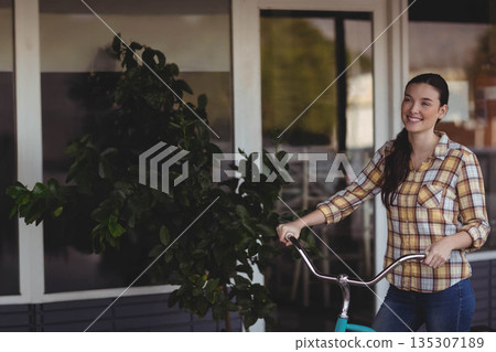 Woman in her twenties holding teal bicycle on tiled patio beside potted plant and glass doors Woman in her twenties holding teal bicycle on tiled patio beside potted plant and glass doors 135307189