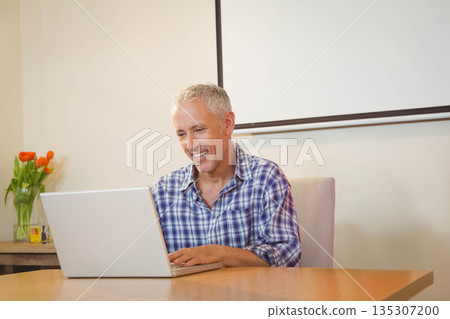 Man wearing plaid shirt typing on laptop at home workspace with wooden table and orange tulips 135307200