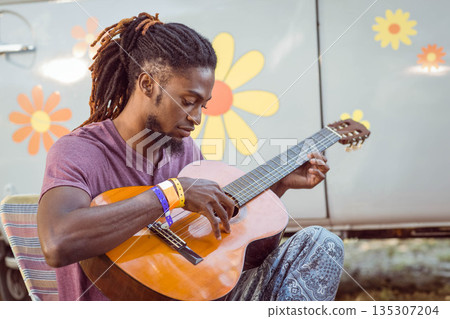 African American man strumming acoustic guitar on striped chair outdoors beside flowered van African American man strumming acoustic guitar on striped chair outdoors beside flowered van 135307204