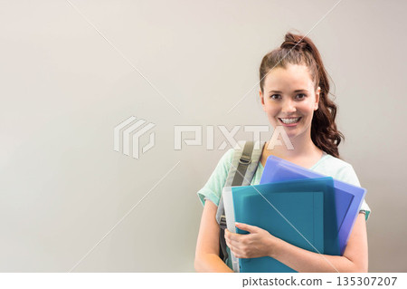 Teenage student standing against pale wall holding blue and teal binders, grey backpack, copy space Teenage student standing against pale wall holding blue and teal binders, grey backpack, copy space 135307207