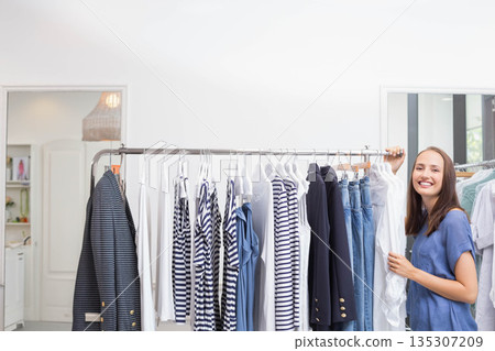 Woman organizing striped tops and denim on metal rack in retail boutique by window, copy space Woman organizing striped tops and denim on metal rack in retail boutique by window, copy space 135307209