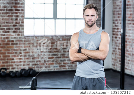 Muscular man in gray sleeveless shirt and dark shorts standing inside gym by dumbbells and barbell 135307212