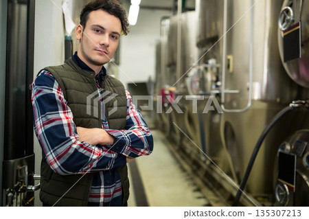 Male brewer wearing green vest and plaid shirt standing in brewery corridor by fermentation tanks Male brewer wearing green vest and plaid shirt standing in brewery corridor by fermentation tanks 135307213