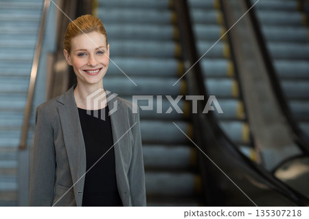 Businesswoman standing and smiling in lobby near escalators with metal handrails, copy space 135307218