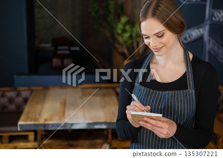 Female server standing beside table at restaurant writing order on notepad with pen, copy space 135307221