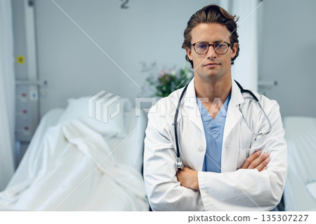 Male doctor standing with arms crossed next to hospital bed in scrubs, lab coat with stethoscope Male doctor standing with arms crossed next to hospital bed in scrubs, lab coat with stethoscope 135307227