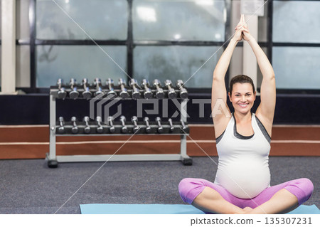 Pregnant woman raising arms overhead, sitting on blue yoga mat in gym with dumbbells, copy space 135307231