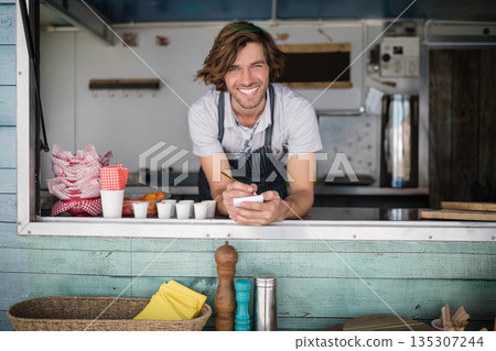 Man leaning forward at street-side food truck window while holding notepad and pen near red napkins Man leaning forward at street-side food truck window while holding notepad and pen near red napkins 135307244