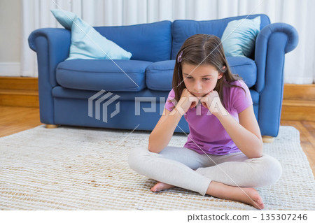 Female child sitting cross-legged on rug in living room near blue sofa, pillows, resting chin 135307246