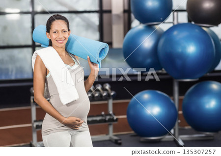 Pregnant woman standing in gym wearing sportswear holding blue yoga mat near dumbbell rack Pregnant woman standing in gym wearing sportswear holding blue yoga mat near dumbbell rack 135307257