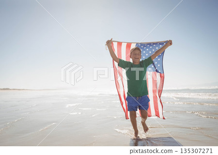 Boy running through shallow ocean water on beach holding American flag billowing under clear sky 135307271