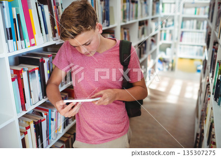 Teenage boy reading paperback book by white bookshelves in aisle with dark backpack, copy space Teenage boy reading paperback book by white bookshelves in aisle with dark backpack, copy space 135307275