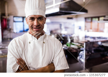 Mature chef standing in restaurant kitchen wearing uniform and toque with stainless steel counters 135307277