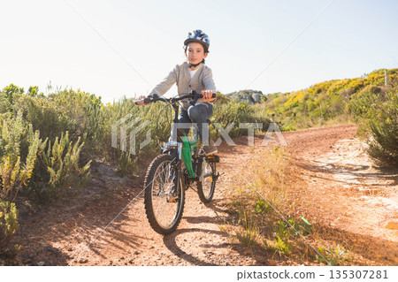 Boy wearing safety helmet riding green mountain bike on rural dirt trail under bright sunlight 135307281