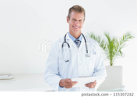Mature male doctor in lab coat smiling while holding tablet in clinic with stethoscope and plant 135307282