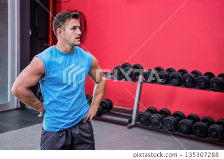 Muscular male posing hands on hips in gym featuring dumbbell rack and glass door, copy space Muscular male posing hands on hips in gym featuring dumbbell rack and glass door, copy space 135307288