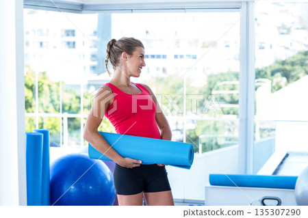 Woman holding rolled blue exercise mat in bright fitness studio with foam rollers and kettlebell 135307290