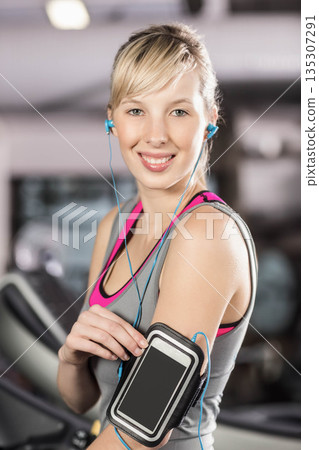 Woman adjusting smartphone armband on upper arm while wearing earphones at gym with cardio machines 135307291