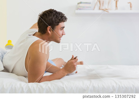 Man lying on bed eating from bowl with spoon in bedroom, shelf holding books, copy space Man lying on bed eating from bowl with spoon in bedroom, shelf holding books, copy space 135307292