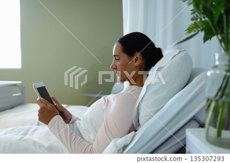 Woman reclining against pillows on bed in bedroom while holding tablet near glass vase greenery Woman reclining against pillows on bed in bedroom while holding tablet near glass vase greenery 135307293