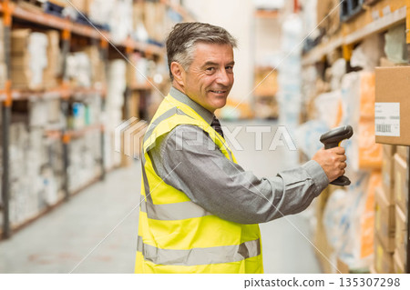 Male warehouse worker scanning boxes with handheld barcode scanner inside aisle wearing safety vest 135307298