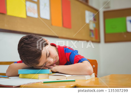 School-age boy sleeping on textbook stack at classroom desk with pencil and open workbook 135307299