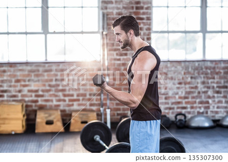 Male lifting hexagonal dumbbell and curling biceps while wearing athletic shorts in gym, copy space 135307300