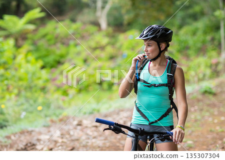 Female cyclist pausing on forest trail and sipping water from tube beside mountain bike, copy space Female cyclist pausing on forest trail and sipping water from tube beside mountain bike, copy space 135307304