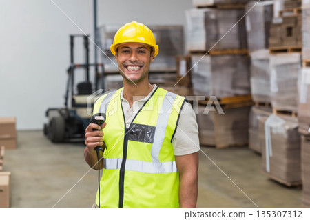 Male warehouse worker in yellow helmet and neon vest scanning boxes with barcode scanner in storage 135307312