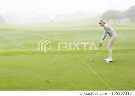 Senior woman lining up putt by flag on dew-covered green with putter and ball, copy space Senior woman lining up putt by flag on dew-covered green with putter and ball, copy space 135307313