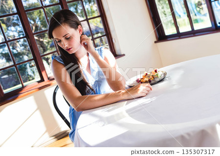 Woman sitting at round white cloth table by wooden windows, sampling pastries and fruit, copy space 135307317