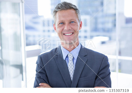 Mature man standing with crossed arms in office by glass windows wearing suit jacket, patterned tie 135307319