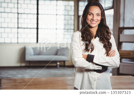 Woman standing with arms crossed and smiling in loft workspace by sofa, grid window, copy space 135307323