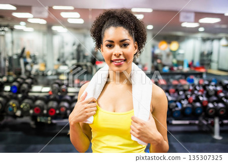 African American woman wearing yellow tank top and holding white towel near dumbbell rack at gym African American woman wearing yellow tank top and holding white towel near dumbbell rack at gym 135307325