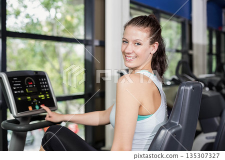 Woman in her twenties adjusting recumbent bike console in sportswear inside gym with treadmills 135307327