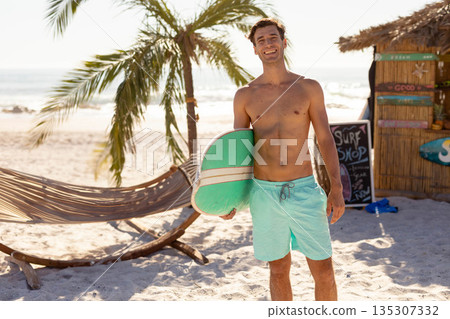 Man holding green-and-white surfboard and wearing turquoise swim trunks on beach under palm trees 135307332